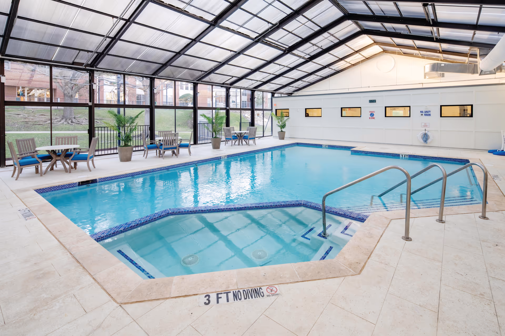 Indoor swimming pool area with clear blue water, surrounded by beige tiled flooring. There are metal handrails leading into the pool and several small tables with chairs and potted plants along the glass wall. The ceiling is transparent, allowing natural light to illuminate the space.