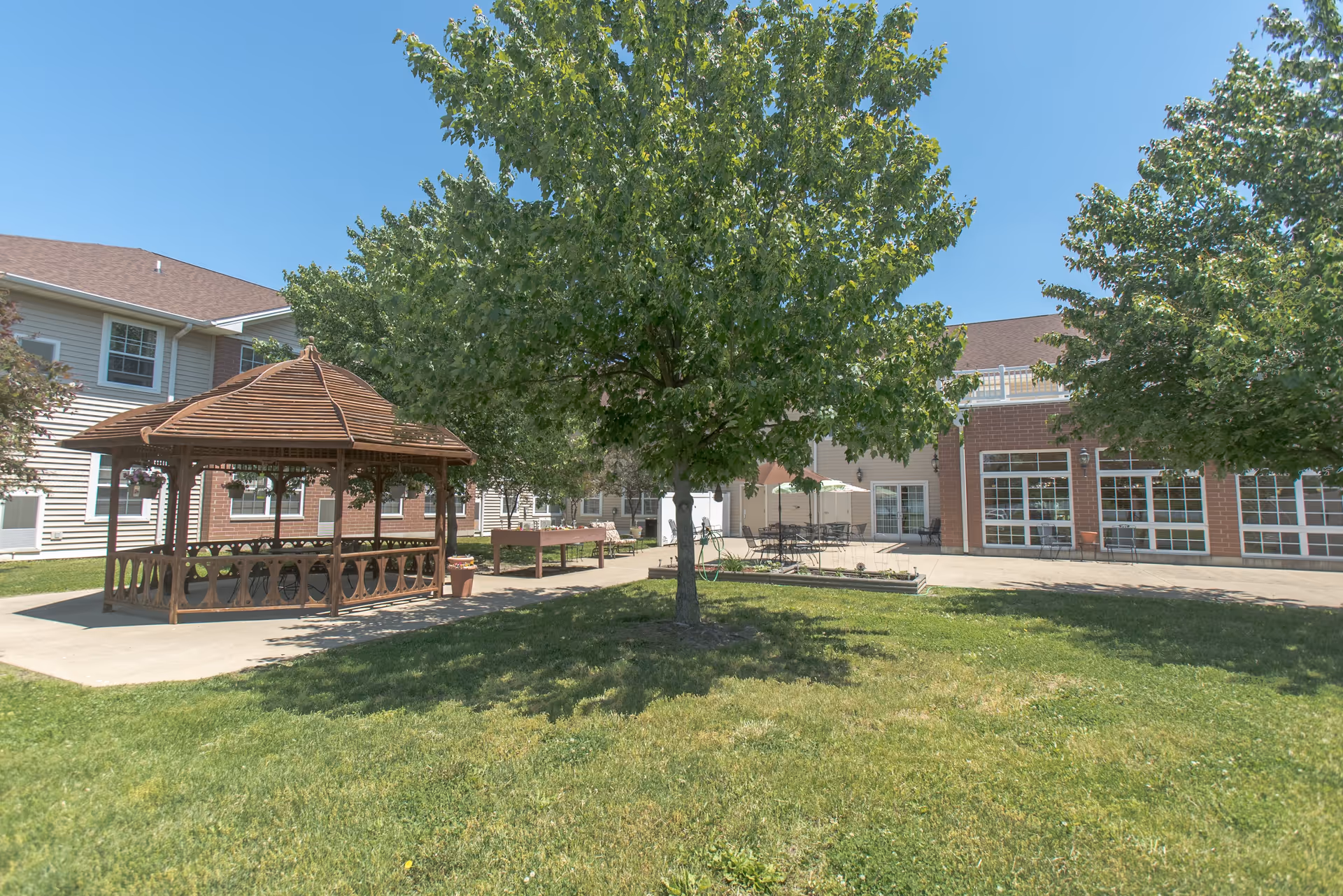 Outdoor area of Bowman Estates of Danville featuring a wooden gazebo, green grass, several trees, and a patio with tables and chairs under umbrellas, adjacent to a two-story building with large windows.