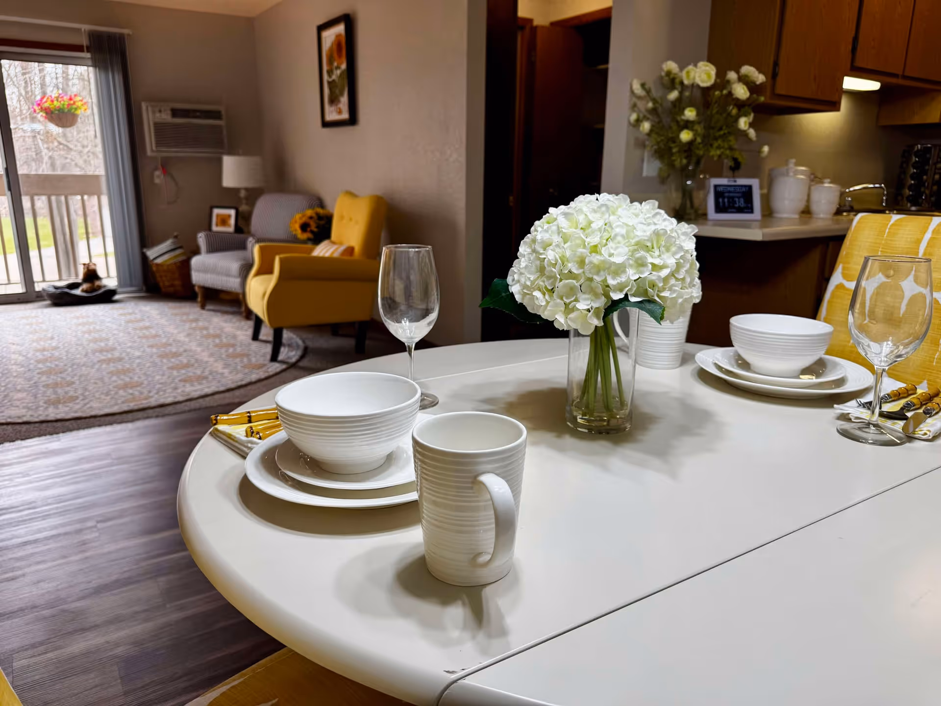 A dining table set for two with white dishes, bowls, mugs, wine glasses, and a vase of white flowers in the center. In the background, there is a living area with a yellow armchair, a gray armchair, a floor lamp, a framed picture on the wall, and a sliding glass door leading to an outdoor balcony with a hanging flower pot.