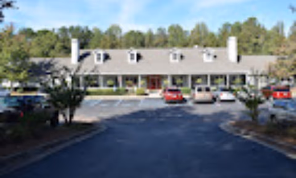 Front exterior view of a single-story assisted living and memory care community building with a gray roof, multiple windows, and a red entrance door. Several cars are parked in the parking lot in front of the building, surrounded by trees and greenery under a clear blue sky.