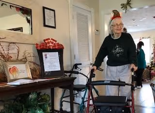 An elderly woman using a walker inside a well-lit room with holiday decorations. There is a table with framed documents and holiday-themed items on the left side, and another person is visible in the background near a doorway.