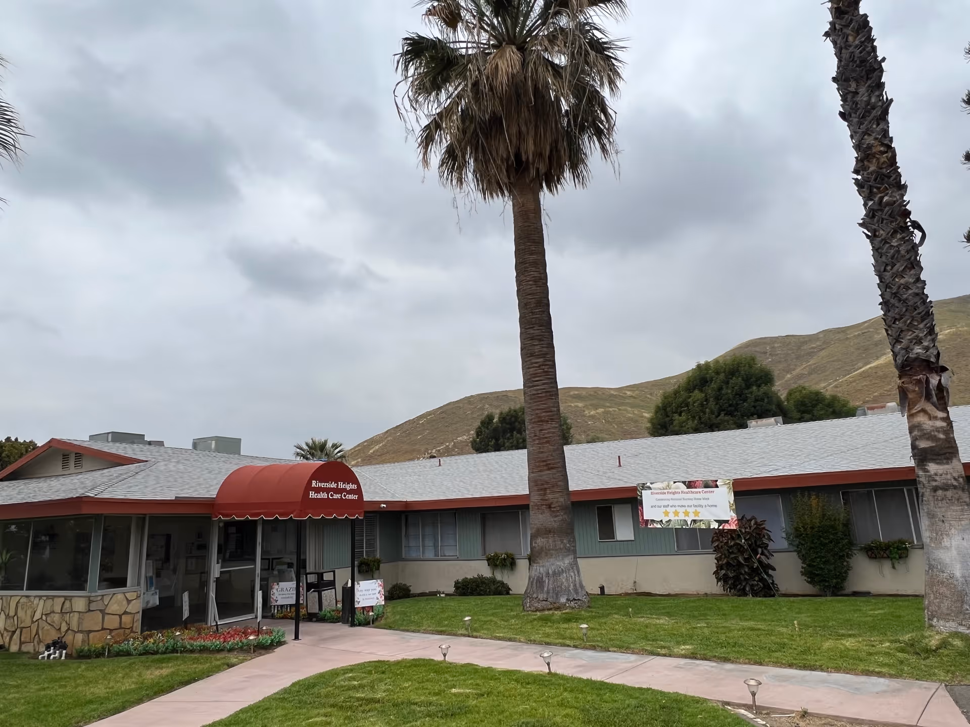 Exterior view of Riverside Heights Healthcare Center, a single-story building with a red awning over the entrance. The building is surrounded by green grass, palm trees, and hills in the background under a cloudy sky.