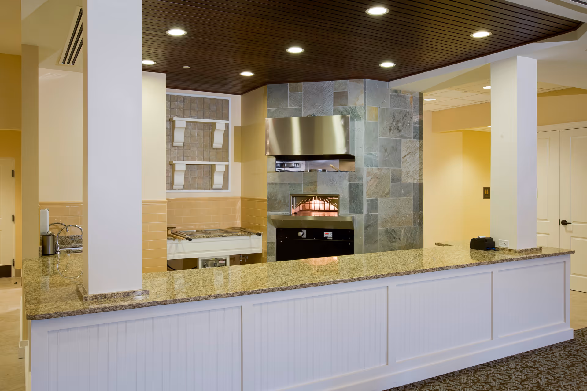 Interior view of a kitchen area with a granite countertop bar, a stone-tiled wall with a stainless steel oven and hood, and a cooking range in the background. The ceiling has recessed lighting and wooden paneling.