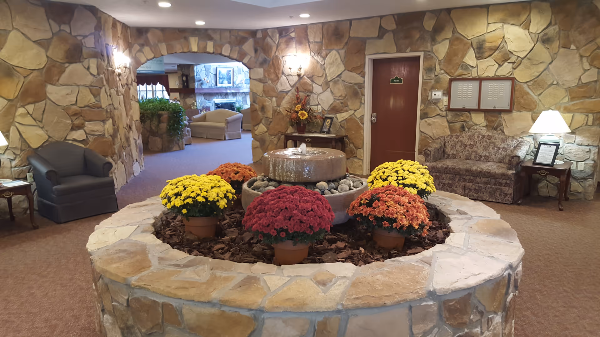 Interior view of a cozy common area in a senior living facility with stone walls and a circular stone planter in the center containing colorful potted flowers and a small water fountain. There are armchairs and a sofa around the room, a table with a lamp, and a doorway leading to another room with more seating and a fireplace.