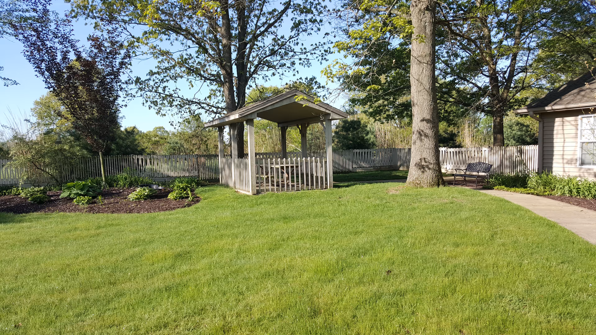 A well-maintained outdoor garden area with green grass, a wooden gazebo with a picnic table inside, a large tree, a bench, and a paved walkway. The area is surrounded by a wooden fence and various plants and shrubs.