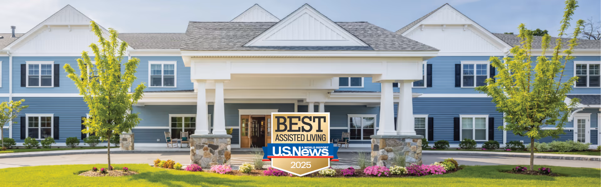 Exterior view of a large, two-story assisted living facility with blue siding and white trim. The entrance features a covered portico supported by white columns with stone bases. There are landscaped flower beds with pink flowers and small trees in front of the building. A sign in front of the entrance reads 'Best Assisted Living U.S. News 2025'.