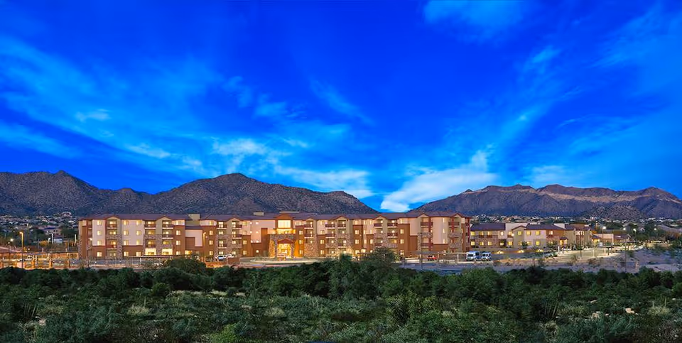 Wide exterior view of Canyon Winds Assisted Living & Memory Care facility at dusk with mountains in the background and a vibrant blue sky with clouds above.