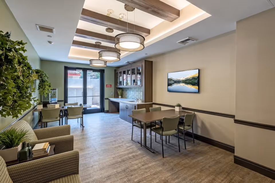 A modern dining room with two tables and several chairs, a wall-mounted TV displaying a scenic mountain and lake view, green walls with decorative plants, and a ceiling with wooden beams and round pendant lights. There is a door leading outside at the far end of the room.
