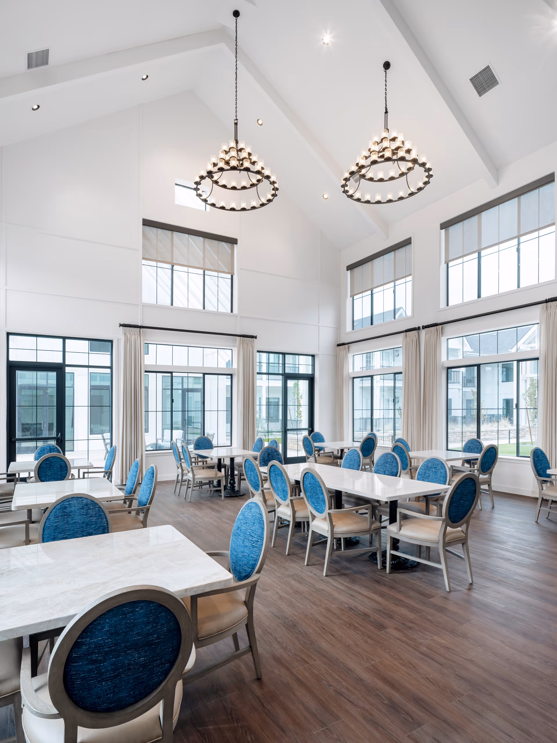 Bright and spacious dining room with high vaulted ceilings, large windows with white curtains, and multiple tables with blue cushioned chairs arranged neatly on a wooden floor. Two large circular chandeliers hang from the ceiling.