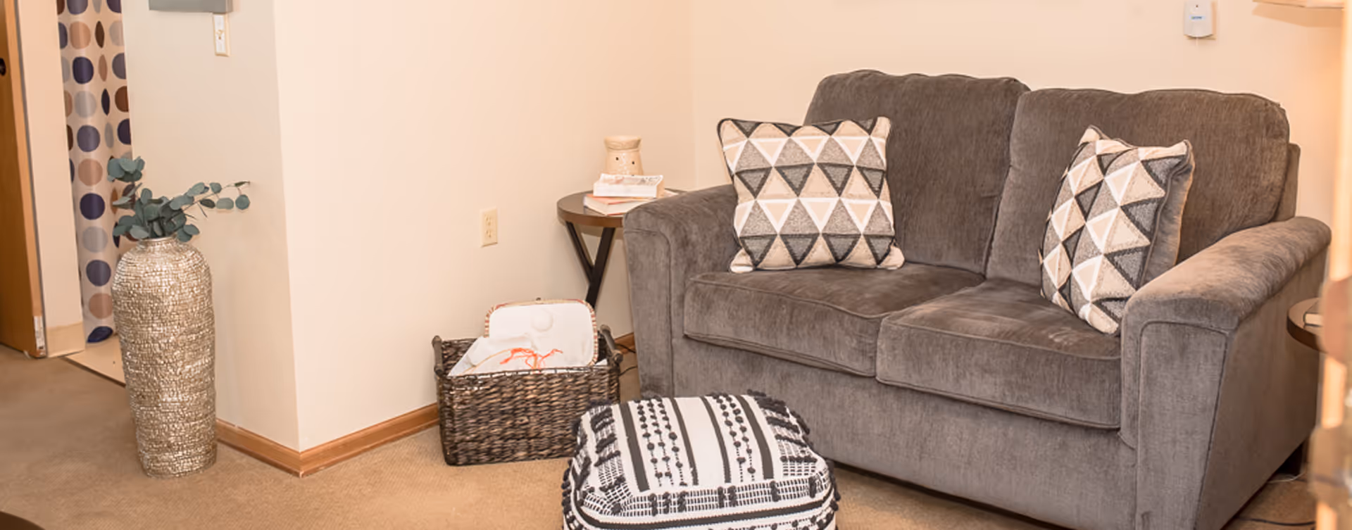A cozy living room area with a gray upholstered loveseat featuring two geometric patterned pillows. In front of the loveseat is a black and white patterned ottoman. To the left, there is a woven basket with some items inside and a small round side table with a candle and books. A tall decorative vase with greenery is placed near the corner, and a doorway with a curtain featuring large polka dots is visible in the background.