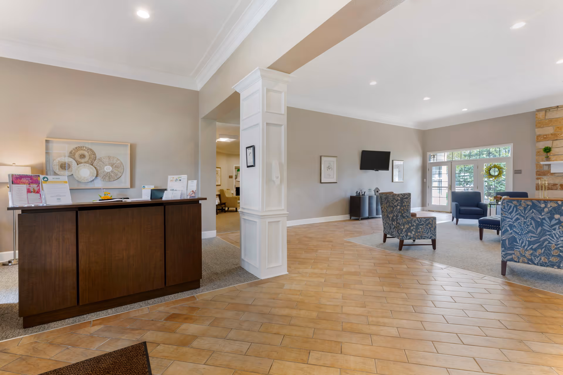 A spacious senior living facility lobby with a dark wooden reception desk on the left, a white decorative column in the center, and a seating area with patterned and solid blue chairs near a stone fireplace and large windows letting in natural light.