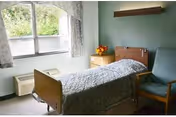 A small nursing facility bedroom with a single bed covered in a patterned quilt, a wooden nightstand with a vase of flowers, a green upholstered chair, and a window with floral curtains letting in natural light.