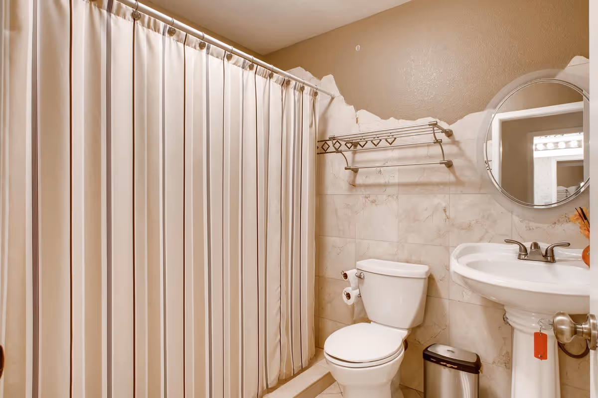 A bathroom with a white toilet, a pedestal sink with a silver faucet, a round mirror above the sink, a metal towel rack mounted on the wall, a striped shower curtain, and a small trash can on the floor.
