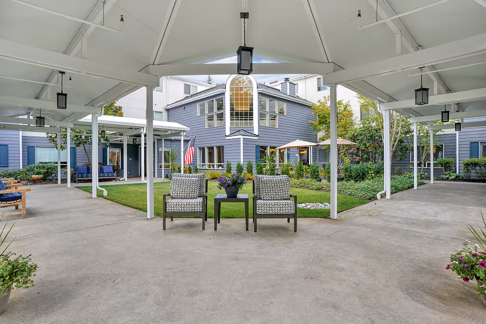 Covered outdoor seating area with two patterned chairs and a small table with a flower pot in the center, surrounded by a garden and a two-story building with large windows and an American flag.