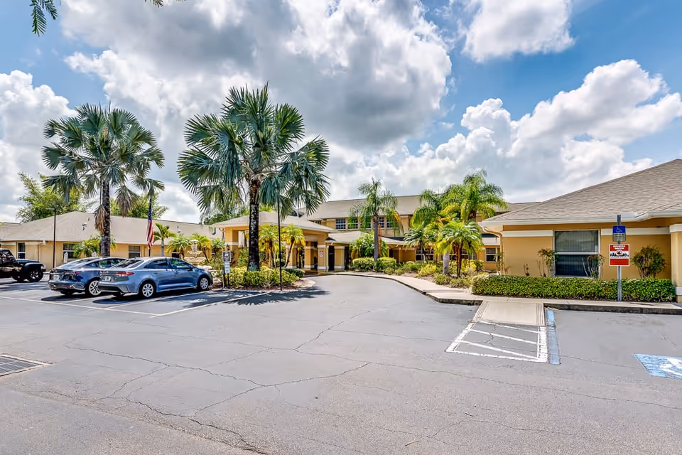 Front entrance of a single-story yellow senior living building with palm trees, parked cars, and a partly cloudy sky.