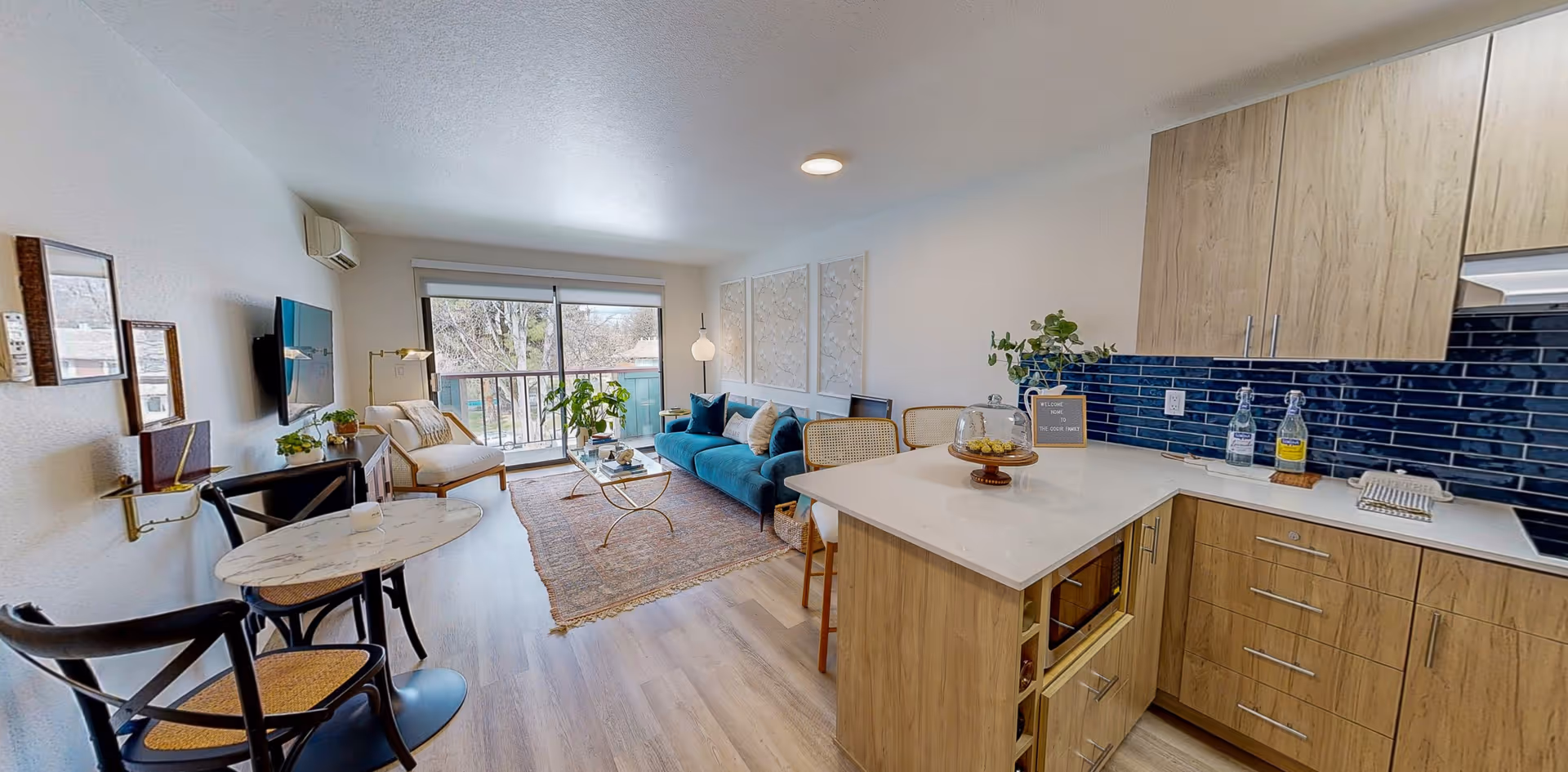 A bright and modern open-concept living space featuring a kitchen with light wood cabinets and a white countertop, a small dining area with a round marble table and two chairs, and a living room with a blue sofa, armchair, coffee table, and large window with a view outside.