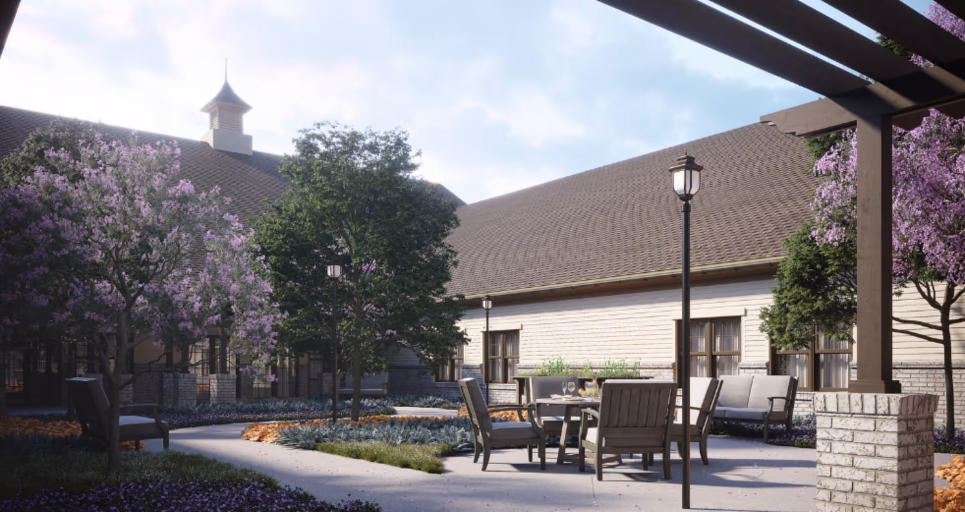 Outdoor courtyard area at Alto Johns Creek featuring a paved walkway, several trees with purple blossoms, outdoor seating with chairs and a table set with glasses, and a building with a sloped roof in the background under a partly cloudy sky.