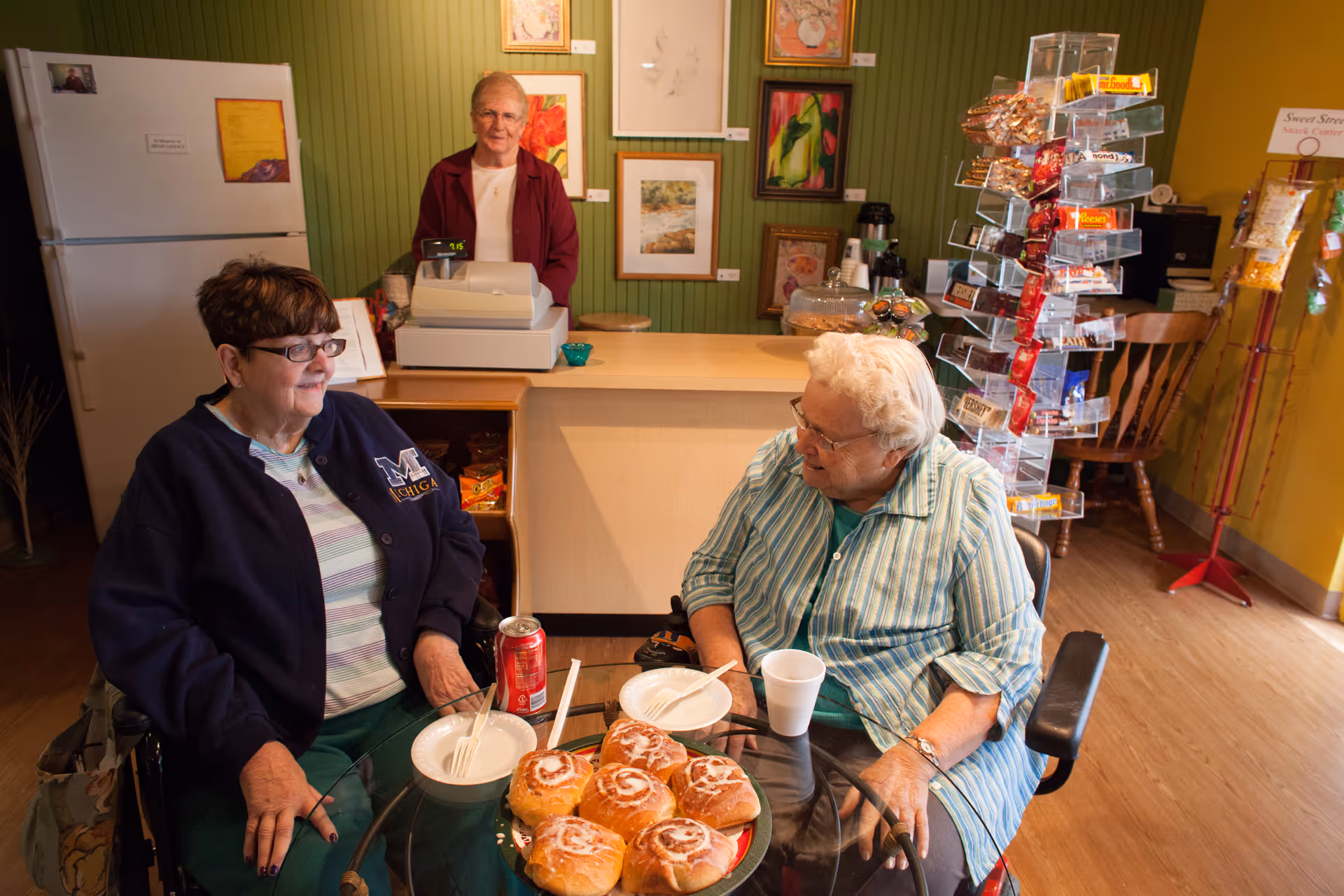 Two elderly women sitting at a round glass table with cinnamon rolls and drinks, engaging in conversation inside a snack center. Behind the counter, a woman stands near a cash register. The room has green and yellow walls with framed artwork and a rotating display rack filled with snacks.