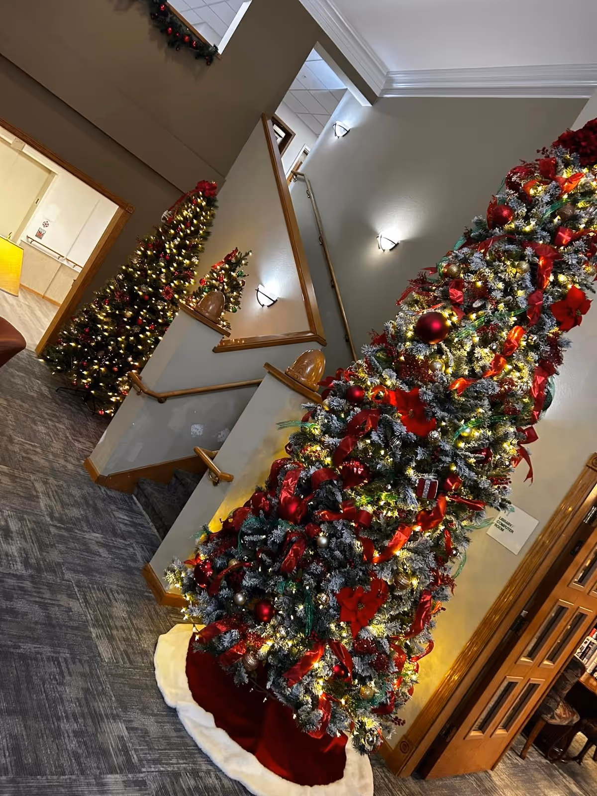 Interior view of a senior living facility decorated for Christmas with two Christmas trees adorned with red ribbons, ornaments, and lights. The scene includes a carpeted floor, a staircase with wooden handrails, wall sconces, and a glimpse into an adjacent room with a yellow lamp.