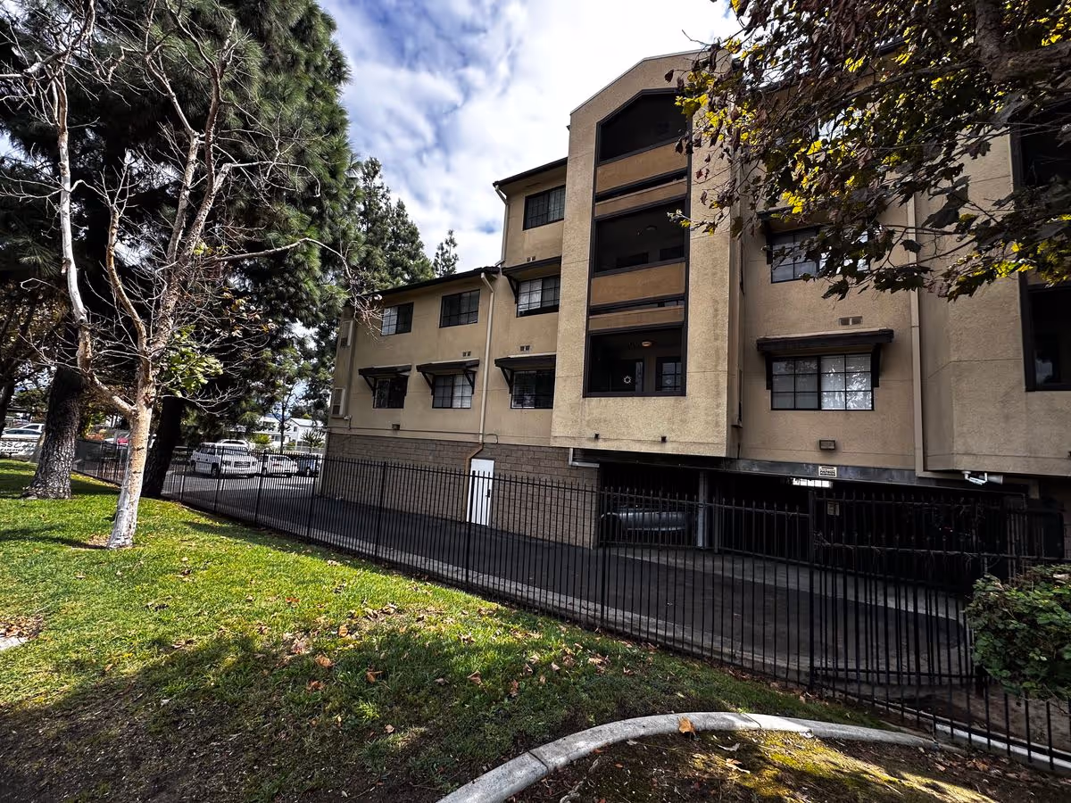Exterior view of a multi-story beige senior living building with gated parking, trees, and a grassy lawn.