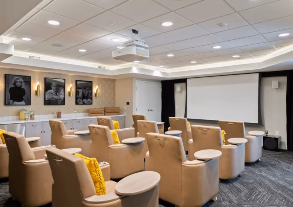A modern media room with multiple beige recliner chairs arranged in rows facing a large projector screen. Each chair has a small attached table and a yellow cushion. The room has a white ceiling with recessed lighting and a mounted projector. The back wall features three black and white framed portraits and a countertop with jars and wooden boxes.