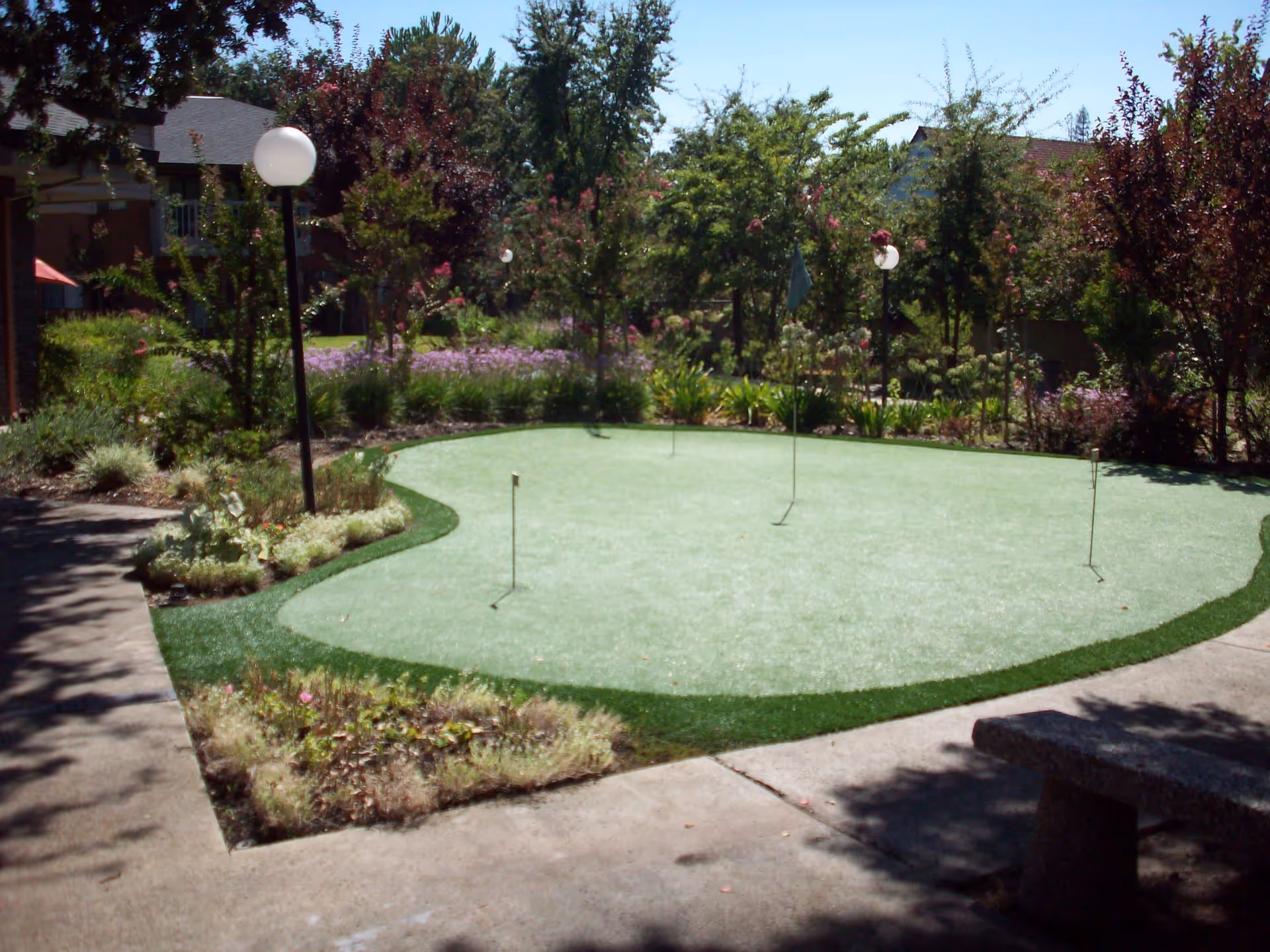 Outdoor putting green surrounded by trees, shrubs, and flowers with a concrete walkway and a stone bench nearby under a sunny sky.