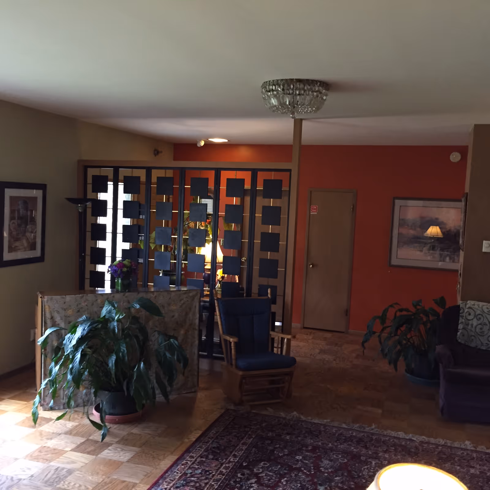 A cozy living room area with wooden parquet flooring and a patterned rug. There is a wooden rocking chair with a blue cushion, several potted plants, and a decorative room divider with square cutouts. The walls are painted in warm tones, including a prominent orange accent wall with a closed wooden door and framed artwork. A ceiling light fixture is visible, and a lamp is reflected in the framed picture on the wall.