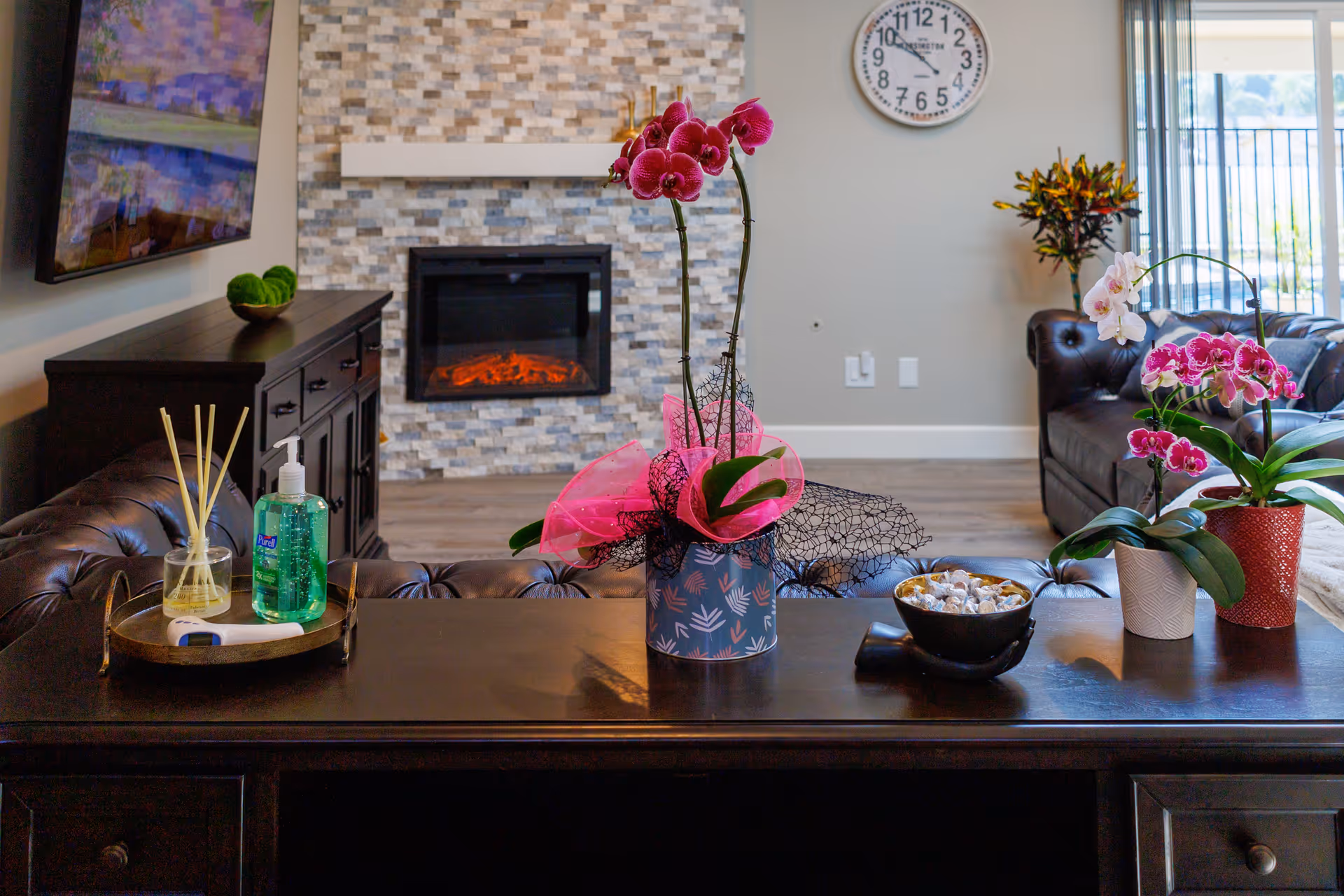 A cozy living room with a dark wooden table in the foreground holding a tray with hand sanitizer, a reed diffuser, a thermometer, and several potted orchids. In the background, there is a stone fireplace with an electric fire, a wall clock above it, a dark leather sofa, and a large window letting in natural light.