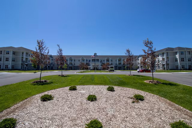 Wide exterior view of a large, three-story senior living facility building with multiple windows and a central entrance. The foreground features a landscaped area with small bushes and young trees, surrounded by a green lawn and paved driveway. The sky is clear and blue.