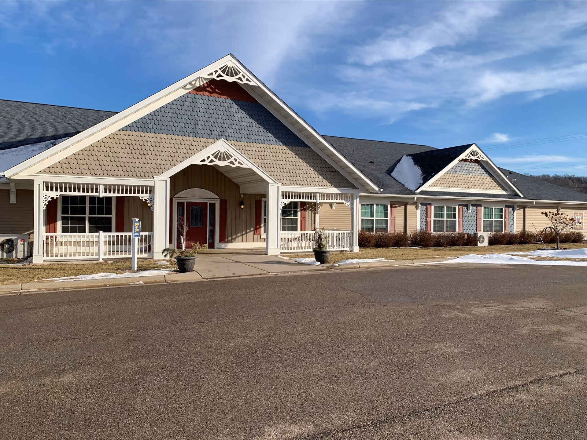 Exterior front view of a single-story senior care facility building with beige siding, decorative gables, and a covered entrance. There are windows with red shutters, a paved driveway, and patches of snow on the ground under a partly cloudy blue sky.
