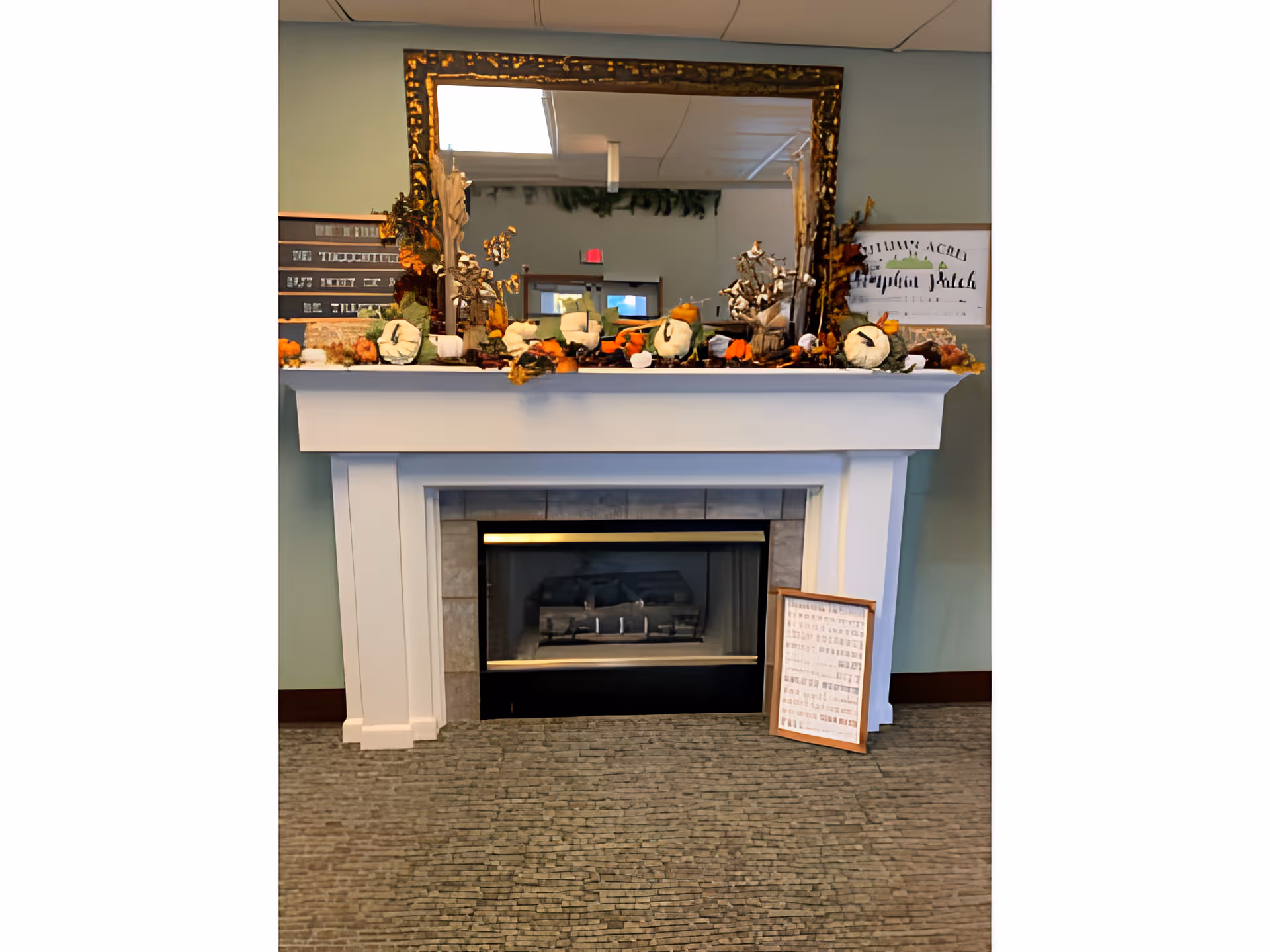 A white fireplace with a tiled interior and a glass front. On the mantel above the fireplace, there are decorative fall-themed items including small pumpkins and leaves. A large ornate gold-framed mirror hangs above the mantel reflecting part of the room. There is a framed sign leaning against the right side of the fireplace. The room has a light blue wall and a carpeted floor.