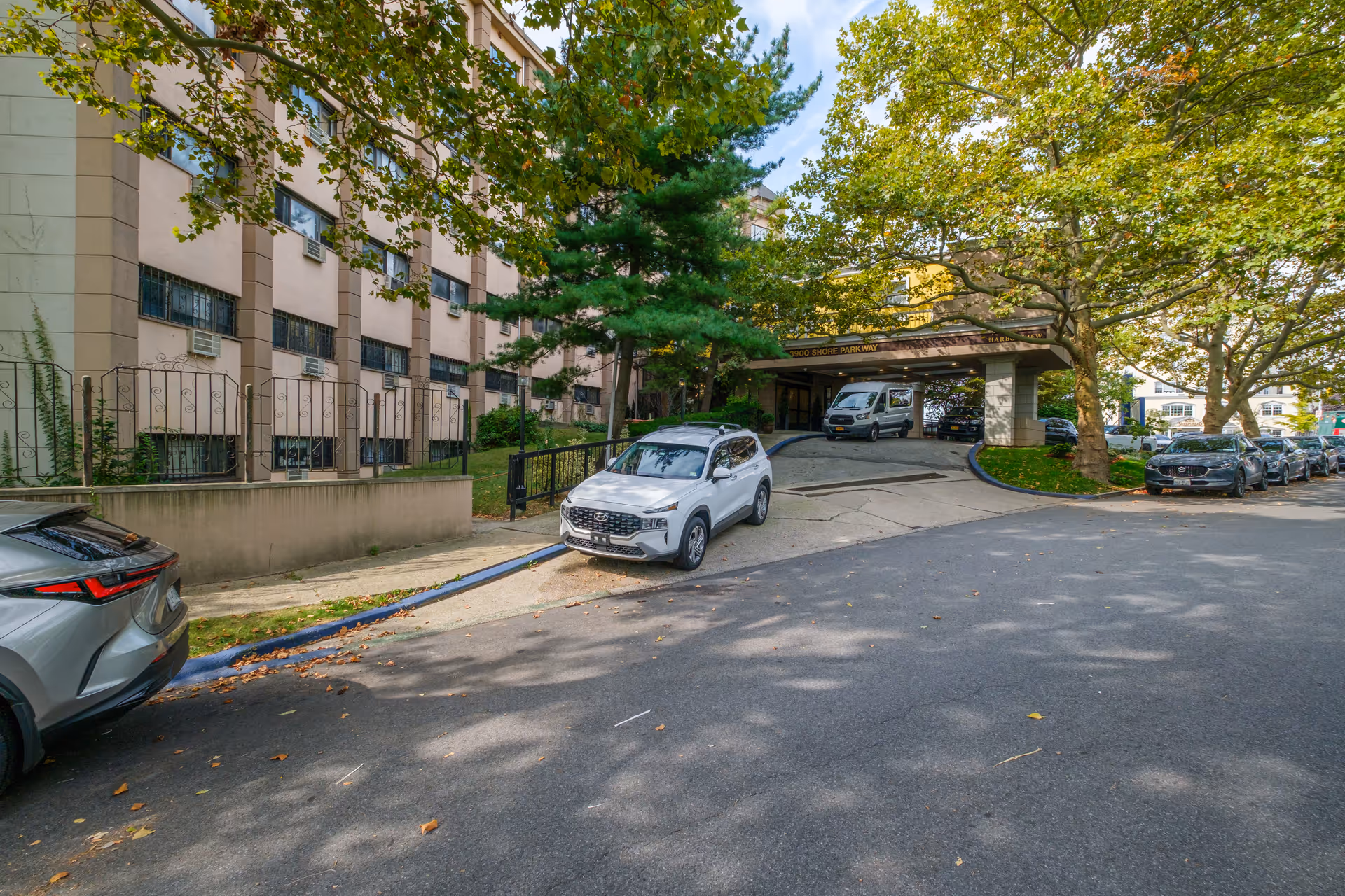 Exterior view of Harbor View Home showing the driveway entrance with several parked cars, trees with green leaves, and a multi-story building with windows and air conditioning units.