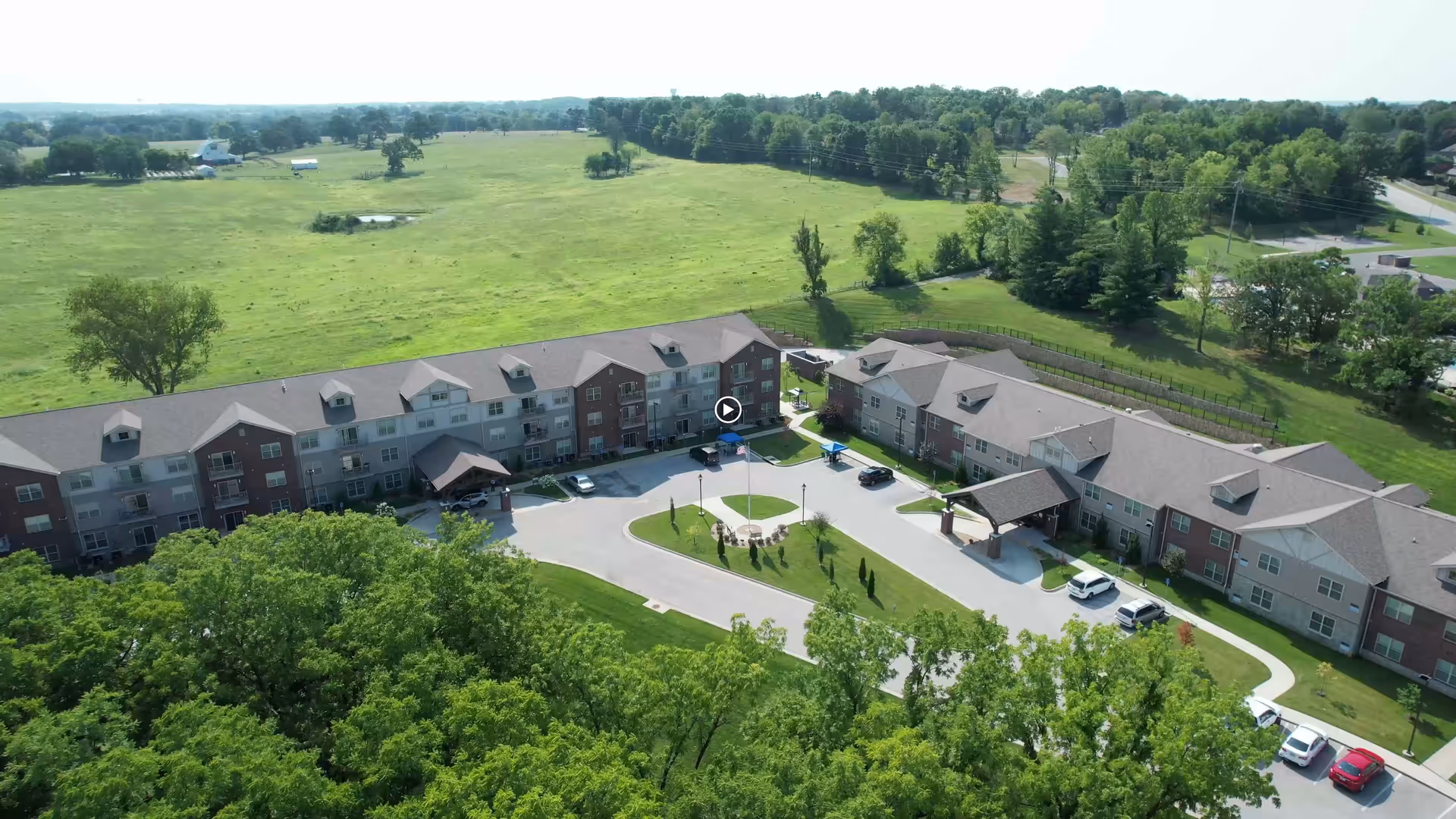Aerial view of The Castlewood Senior Living facility showing two large connected buildings surrounded by green lawns and trees, with a circular driveway and several parked cars. The background features open fields and scattered trees under a clear sky.