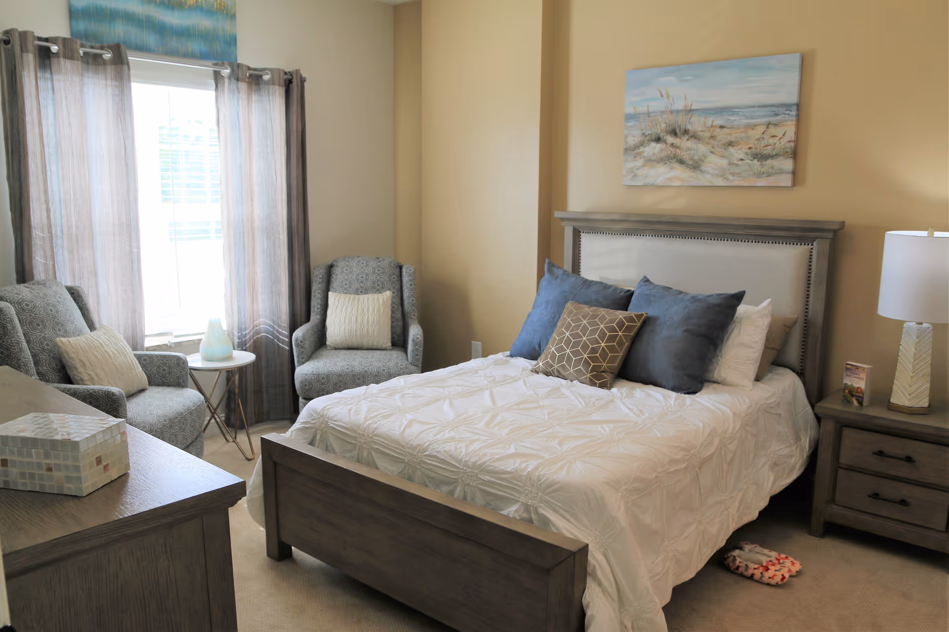 A cozy bedroom with a wooden bed frame and a white comforter, decorated with blue and brown pillows. There are two patterned armchairs with white cushions near a window with sheer gray curtains. A nightstand with a lamp and a small book is beside the bed, and a dresser with a decorative box is in the foreground. A painting of a beach scene hangs on the beige wall above the bed.