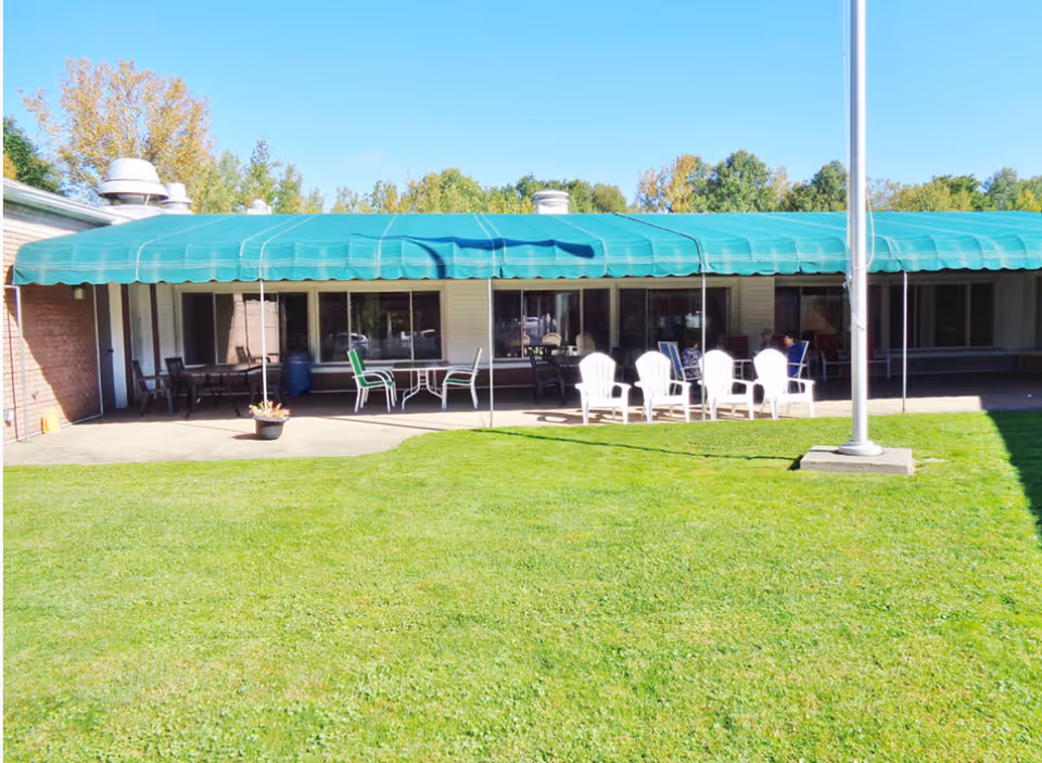 Outdoor patio area at Betsy Ross Nursing Facility with green awning covering several tables and chairs. White plastic chairs are arranged around a table on the concrete patio, with a well-maintained grassy lawn in the foreground and trees in the background under a clear blue sky.