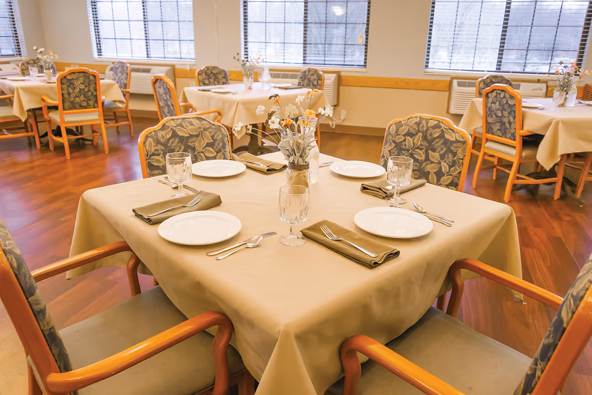 Dining room with a table set for four, floral centerpieces, and several chairs in a senior living facility.