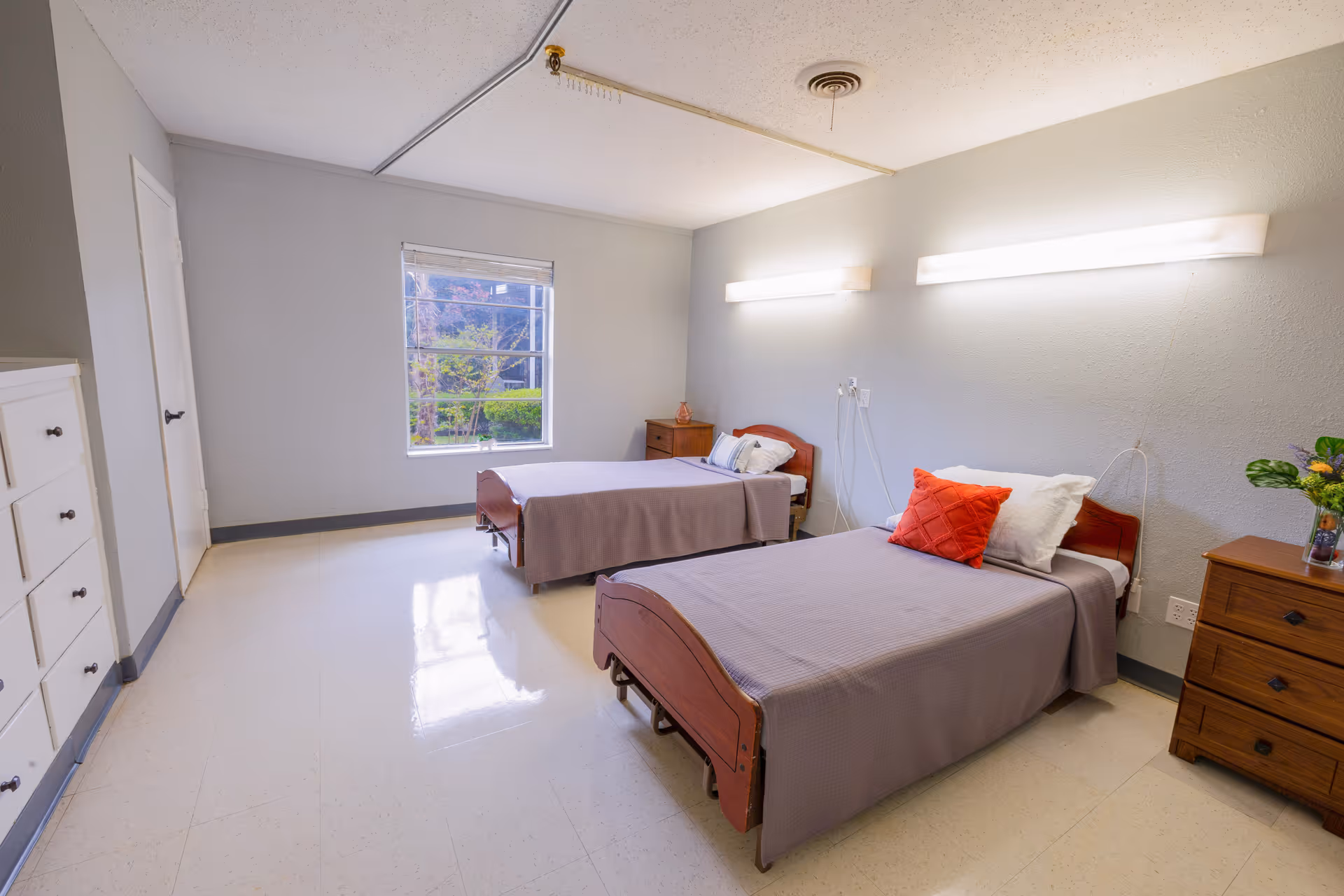 A bright shared nursing facility bedroom with two single beds, bedside tables, and a window showing greenery outside.