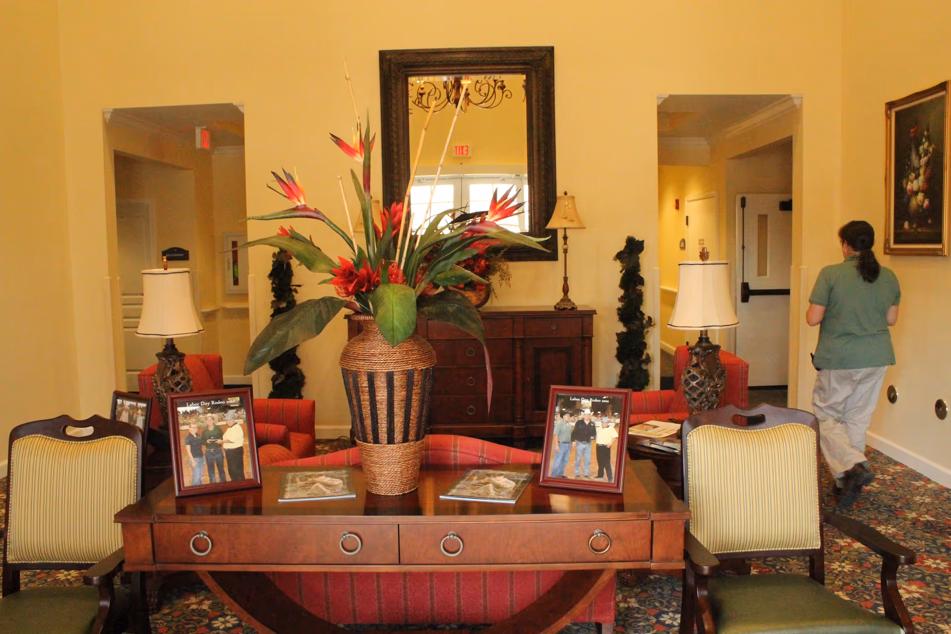 A cozy interior room with a wooden table in the center holding a large decorative vase with tropical flowers and framed photos. Two upholstered chairs with striped cushions are positioned on either side of the table. Behind the table is a wooden sideboard with a lamp and a large mirror hanging above it. A person in a green shirt and light-colored pants is walking away down a hallway on the right side. The room has warm yellow walls and patterned carpet flooring.