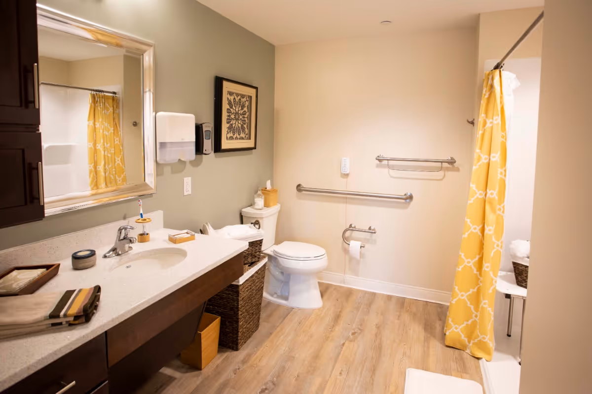 A clean and accessible bathroom with a white toilet, grab bars on the wall, a sink with a large mirror above it, and a shower with a yellow patterned curtain. The floor is wood-style, and there are baskets and towels neatly arranged.