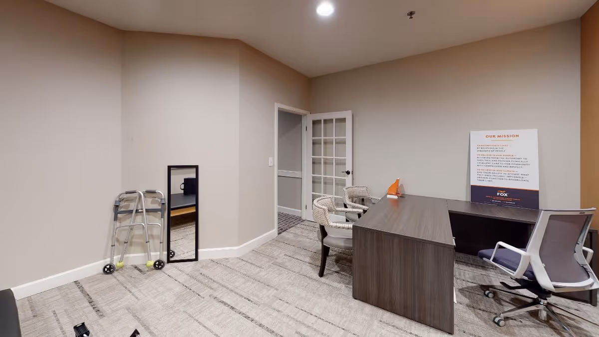 Small interior office/consultation room with a wooden desk, rolling office chair, visitor chairs, a walker, and a floor mirror.