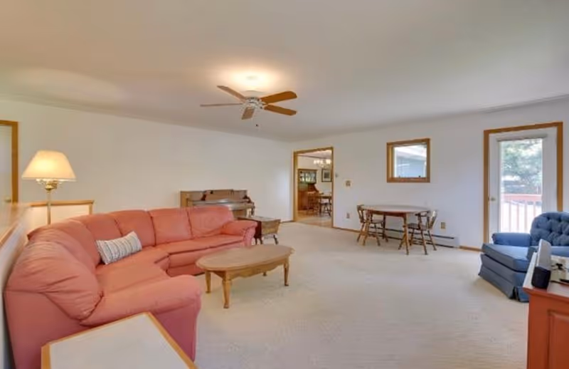 A spacious living room with a large pink sectional sofa, a wooden coffee table, a blue armchair, and a wooden side table with a lamp. There is a ceiling fan above, a piano against the far wall, and a small dining table with two chairs near a window and a glass door leading outside. The room has light-colored carpet and white walls with wooden trim.