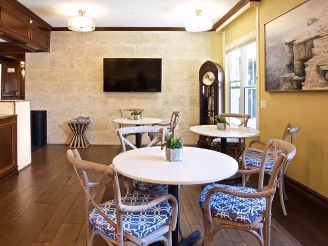 Interior view of a senior living facility dining area with round white tables and wooden chairs with blue patterned cushions. A wall-mounted flat screen TV is visible on a beige stone wall, along with a tall grandfather clock near a window. The room has wooden flooring and warm yellow walls with a framed artwork.
