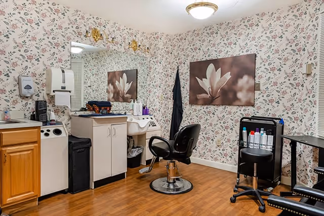 A small salon or grooming room with floral wallpaper, a large mirror, a black salon chair in front of a white sink, a black rolling cart with hair products, and wooden cabinets. The floor is wood, and there is a framed picture of a white flower on the wall.