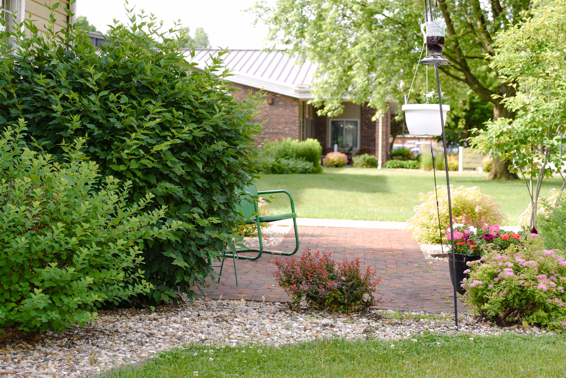 A peaceful outdoor garden area at Maple Crest Campus featuring lush green bushes, a brick patio with green metal chairs, hanging flower pots, and a well-maintained lawn with trees and flowering plants.