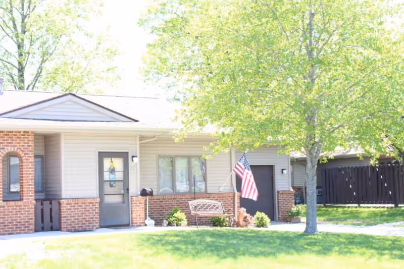 Front exterior of a single-story building with a bench, American flag, tree, and grassy lawn.