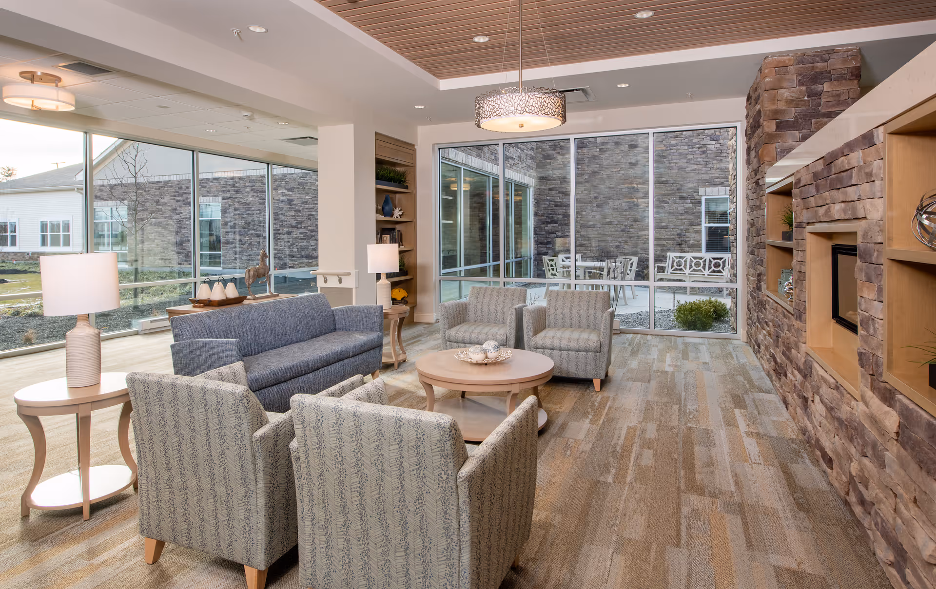 A bright and modern living room area with a blue sofa, four patterned armchairs arranged around a round wooden coffee table, two side tables with lamps, a stone fireplace with built-in shelves, and large windows showing an outdoor patio with seating.