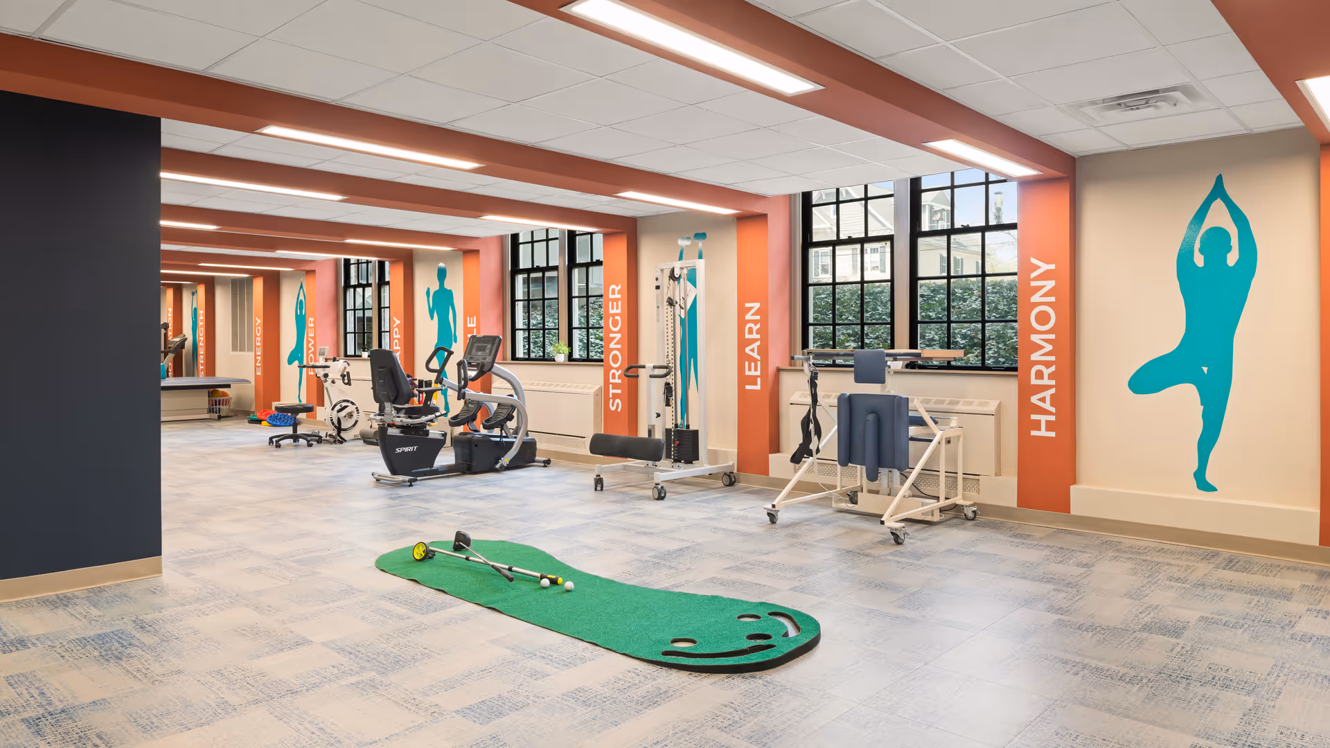 A spacious rehabilitation and fitness room with exercise equipment including stationary bikes, a weight machine, and a putting green mat. The walls feature large windows and motivational words such as ENERGY, STRONGER, LEARN, and HARMONY alongside teal silhouettes of people exercising or stretching.