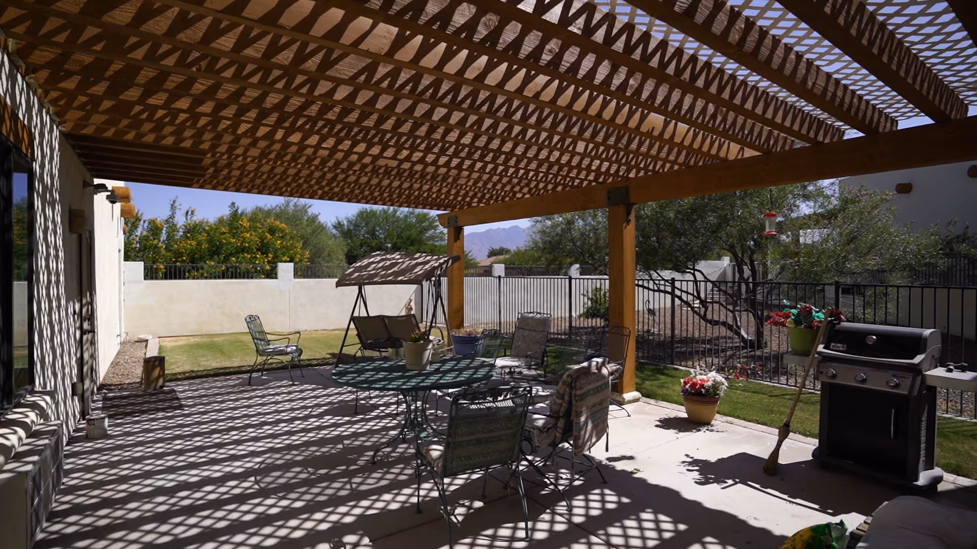 Outdoor patio area with a wooden lattice pergola casting patterned shadows on the concrete floor. The patio is furnished with a round metal table, several chairs with cushions, a swing bench, potted plants, and a barbecue grill. The area is enclosed by a low wall and metal fence, with trees and shrubs visible beyond.