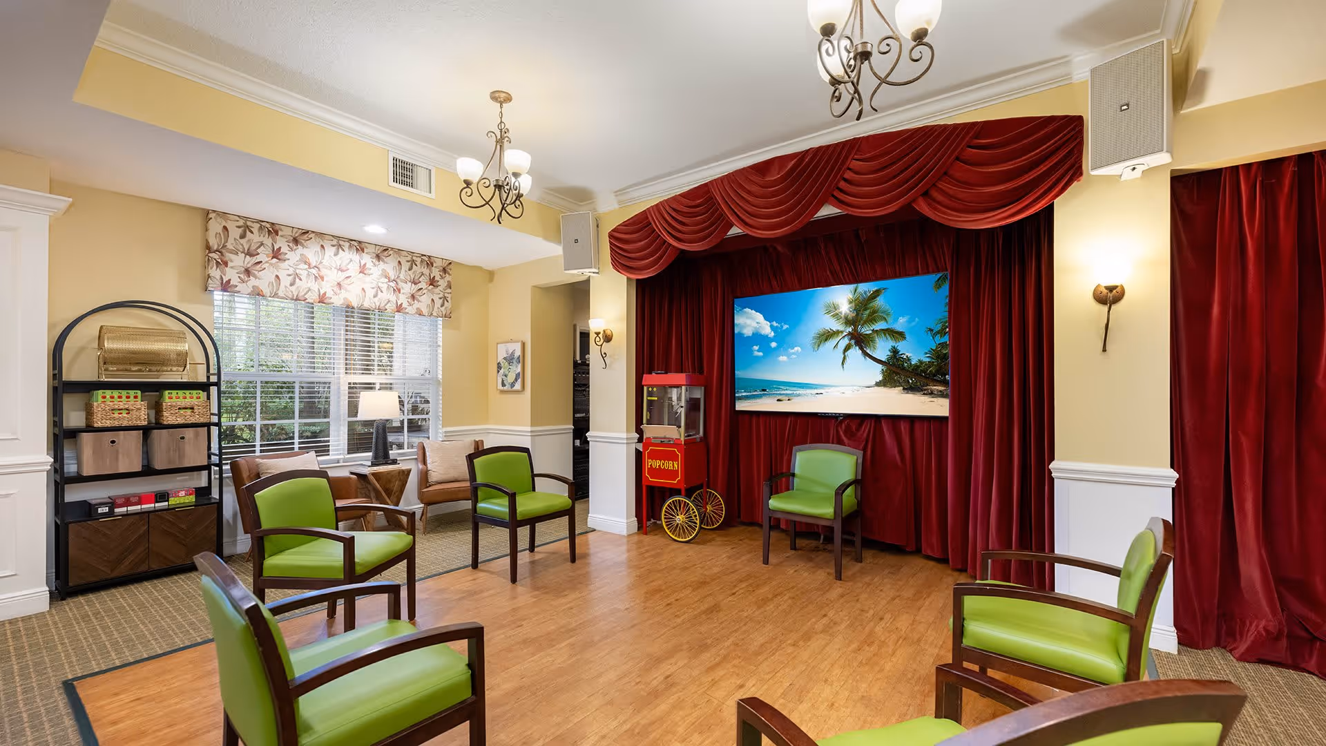 A cozy common area with wooden flooring and green cushioned chairs arranged in a semi-circle facing a small stage with red curtains. On the stage, there is a popcorn machine and a large screen displaying a tropical beach scene. The room has yellow walls, a window with floral curtains, a black metal shelving unit with baskets and boxes, and warm lighting from wall sconces and a ceiling chandelier.