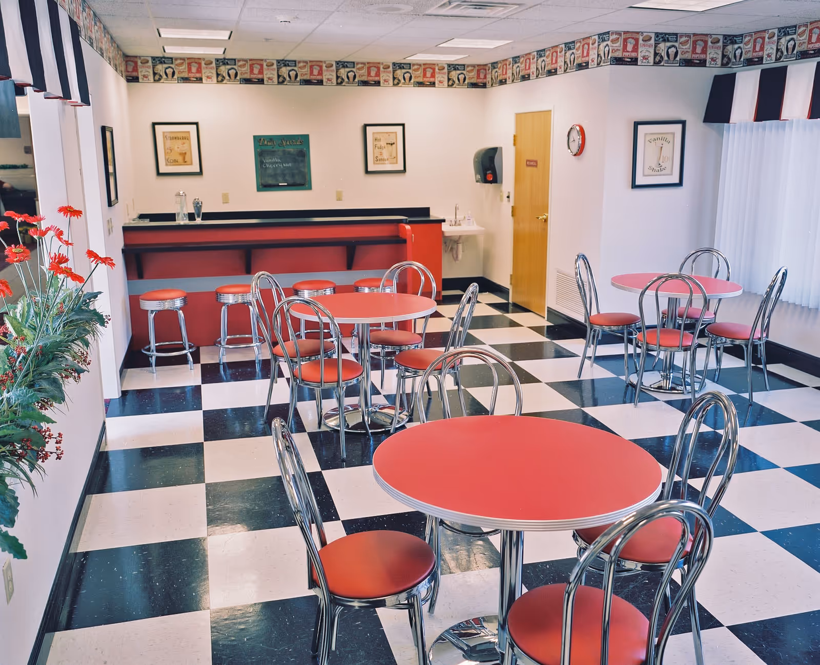Bright retro-style dining room with red round tables, chrome chairs, a black-and-white checkered floor and a counter with stools.
